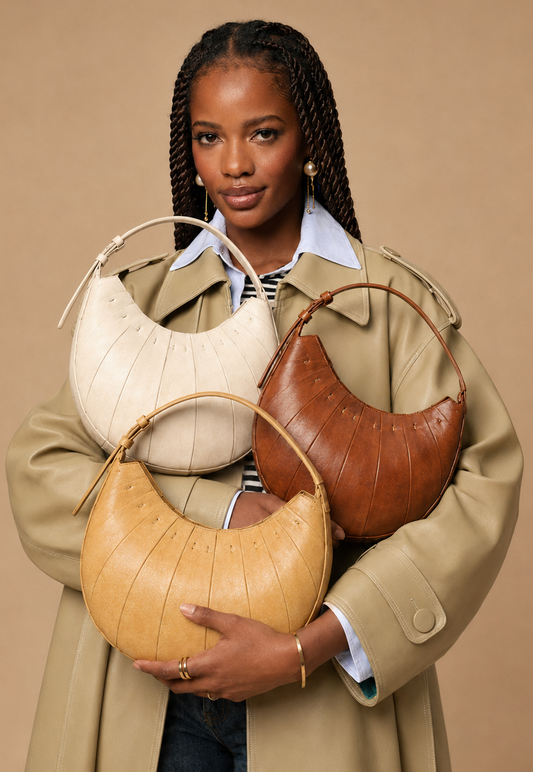 A model wearing a beige trench coat holds three crescent-shaped shoulder bags in cream, tan, and brown against a solid beige background.